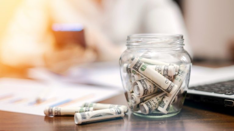 jar-with-rolled-banknotes-table-laptop-papers-woman-background