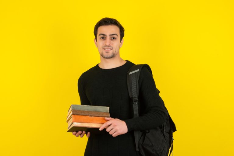portrait-young-male-with-backpack-holding-books-yellow-wall